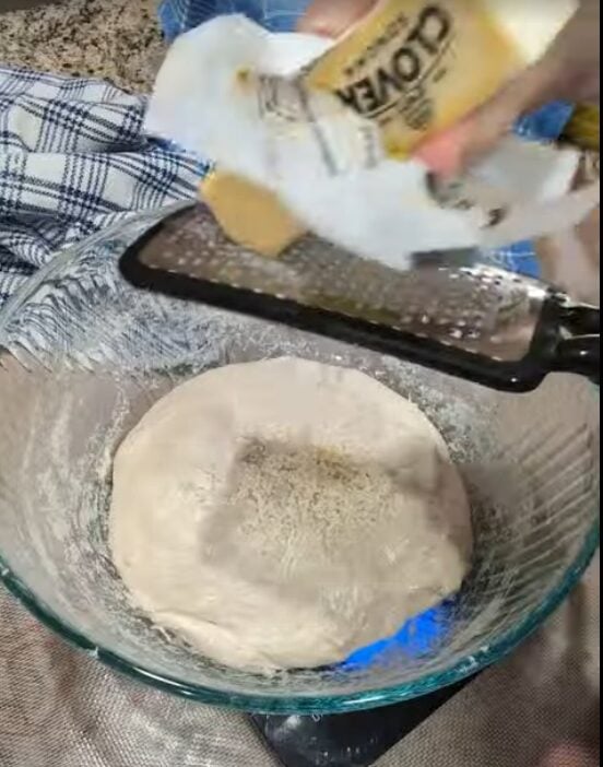 Grating cold butter directly onto sourdough bread roll dough in a glass mixing bowl